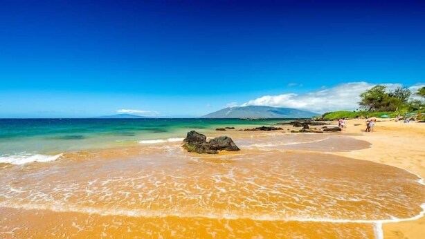 A beach with clear water, a rocky outcrop, and a distant mountain.