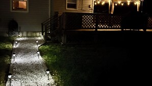 A house with a gravel pathway leading to a porch, a covered area with string lights, and a wooden fence.