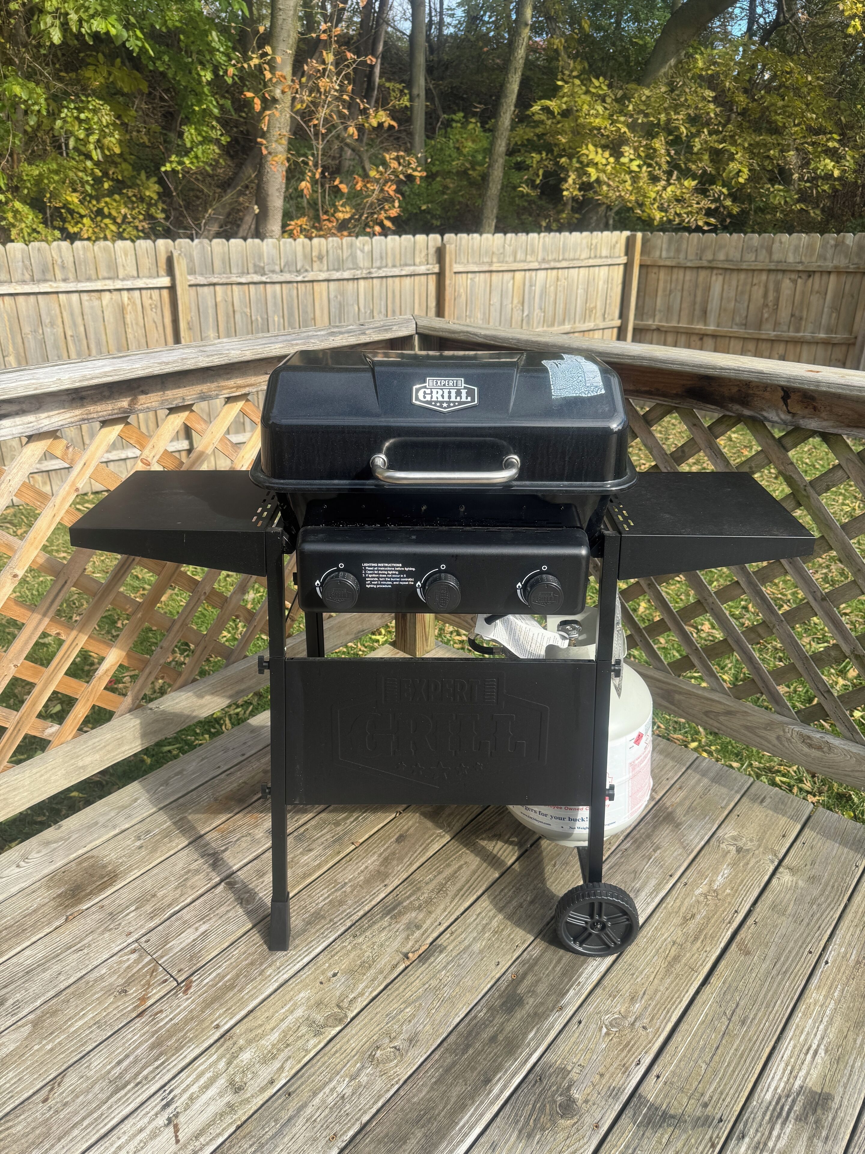 Una parrilla para barbacoa en una terraza de madera, con una valla de madera y árboles al fondo.