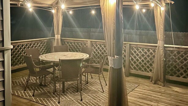 A covered outdoor patio with a round table and chairs, surrounded by a wooden deck and a white fence.