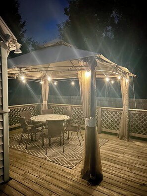 A covered outdoor patio with a round table and chairs, surrounded by a wooden deck and a white fence.