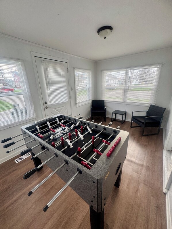 A room with a foosball table, a door, and windows with blinds.
