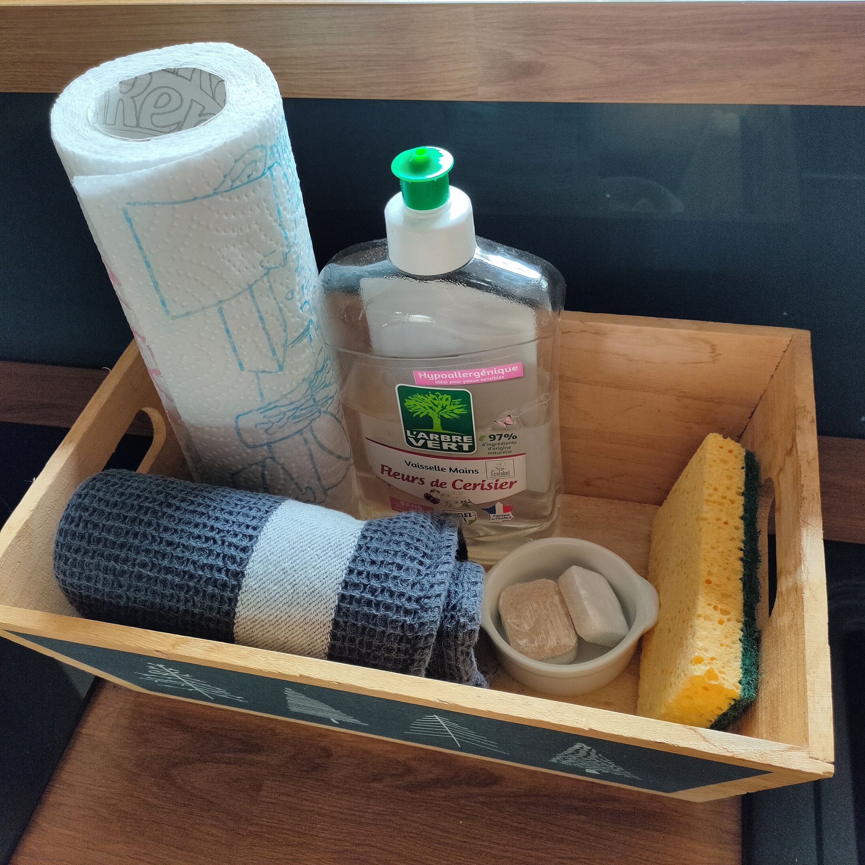 A wooden box containing a roll of toilet paper, a bottle of hand sanitizer, a bar of soap, a sponge, and a knitted item.