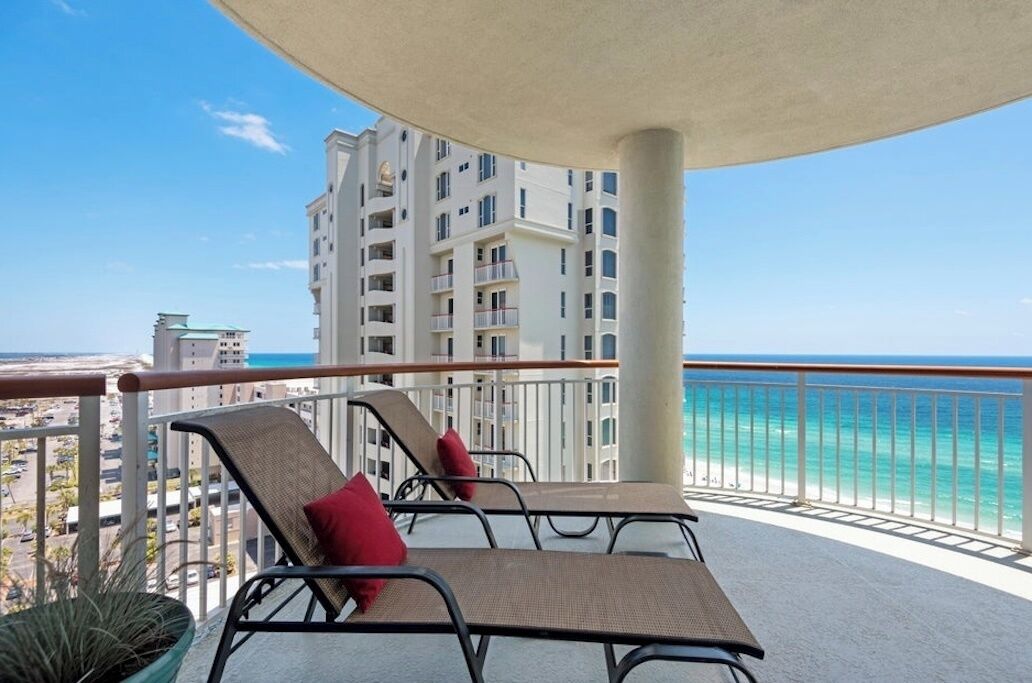A balcony with two lounge chairs and a table, overlooking a beach and a high-rise building.