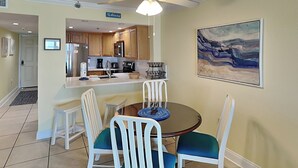 A compact kitchen with wooden cabinets, a microwave, and a sink. There is a dining area with a round table and white chairs. - Ocean Breeze East 602, 2BD/2BA, Gulf-Front, Pool + Private Beach Access (Perdido Key)