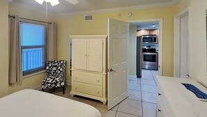 A bedroom with a bed, a ceiling fan, a window with blinds, a chair, a dresser, and an open door leading to a kitchen with an oven and microwave. - Ocean Breeze East 602, 2BD/2BA, Gulf-Front, Pool + Private Beach Access (Perdido Key)