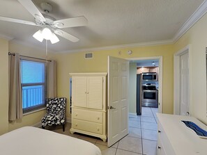 A bedroom with a bed, a ceiling fan, a window with blinds, a chair, a dresser, and an open door leading to a kitchen with an oven and microwave.