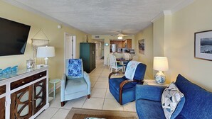 A living room with a TV, a sofa, a chair, and a dining area with a table and chairs. - Ocean Breeze East 602, 2BD/2BA, Gulf-Front, Pool + Private Beach Access (Perdido Key)