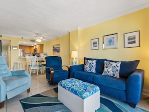 Una sala de estar con un sofá azul, un sillón azul y un otoman a juego. Se ve una cocina al fondo.