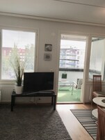 Olohuone, jossa on jalustalla oleva televisio, ruukkukasvi ja näkymä ulkona olevalle parvekkeelle.