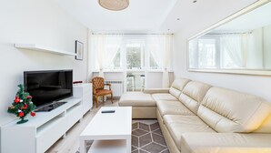Ein modernes Wohnzimmer mit Fernseher, Sofa und Couchtisch. Ein kleiner Weihnachtsbaum und ein Sessel sind ebenfalls vorhanden.