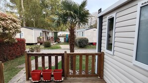 Una terraza de madera con tres macetas rojas, una palmera y una hilera de casas móviles al fondo.