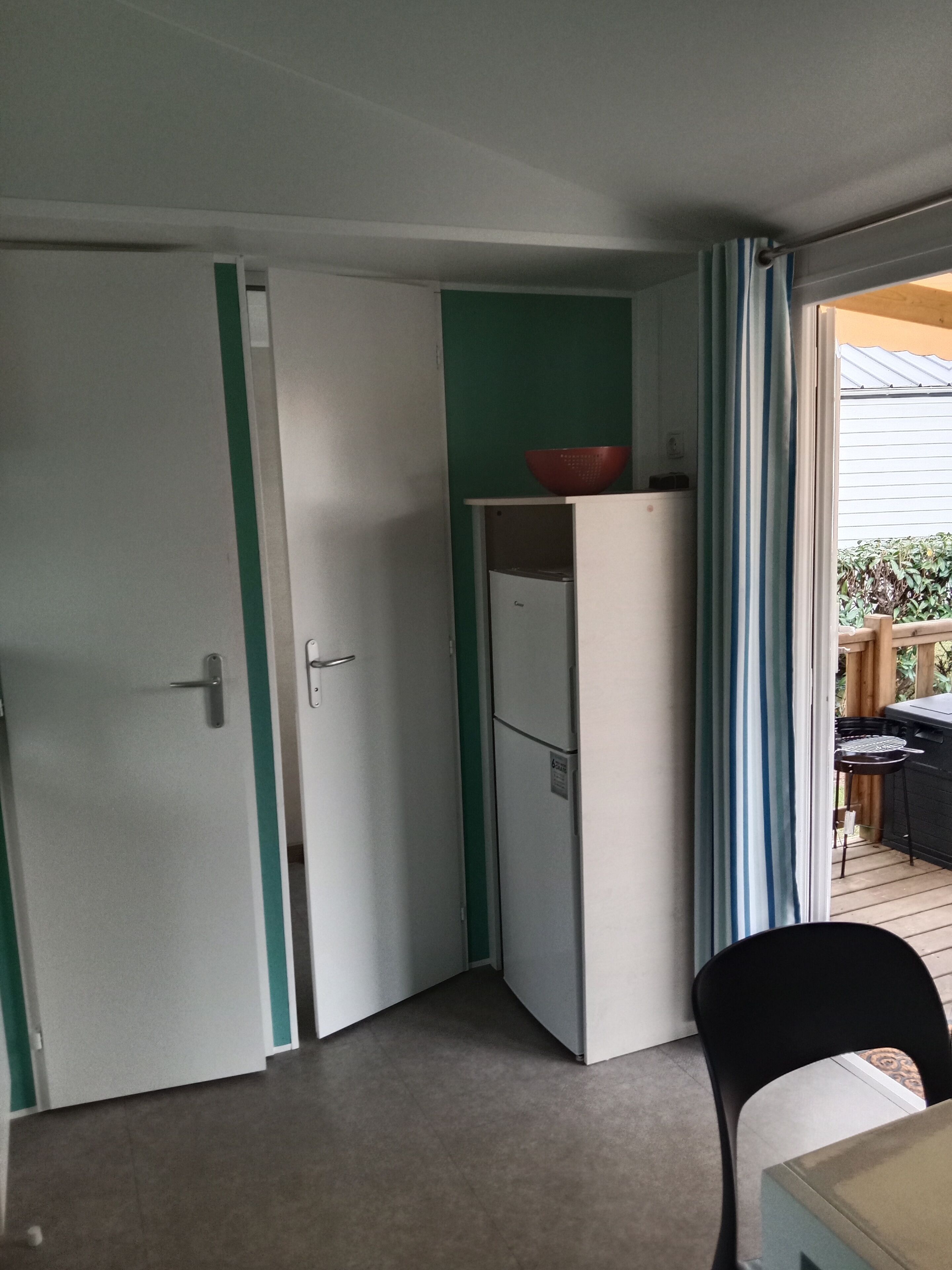 A compact room with a white refrigerator, a door with a green stripe, and a view of a wooden deck outside.