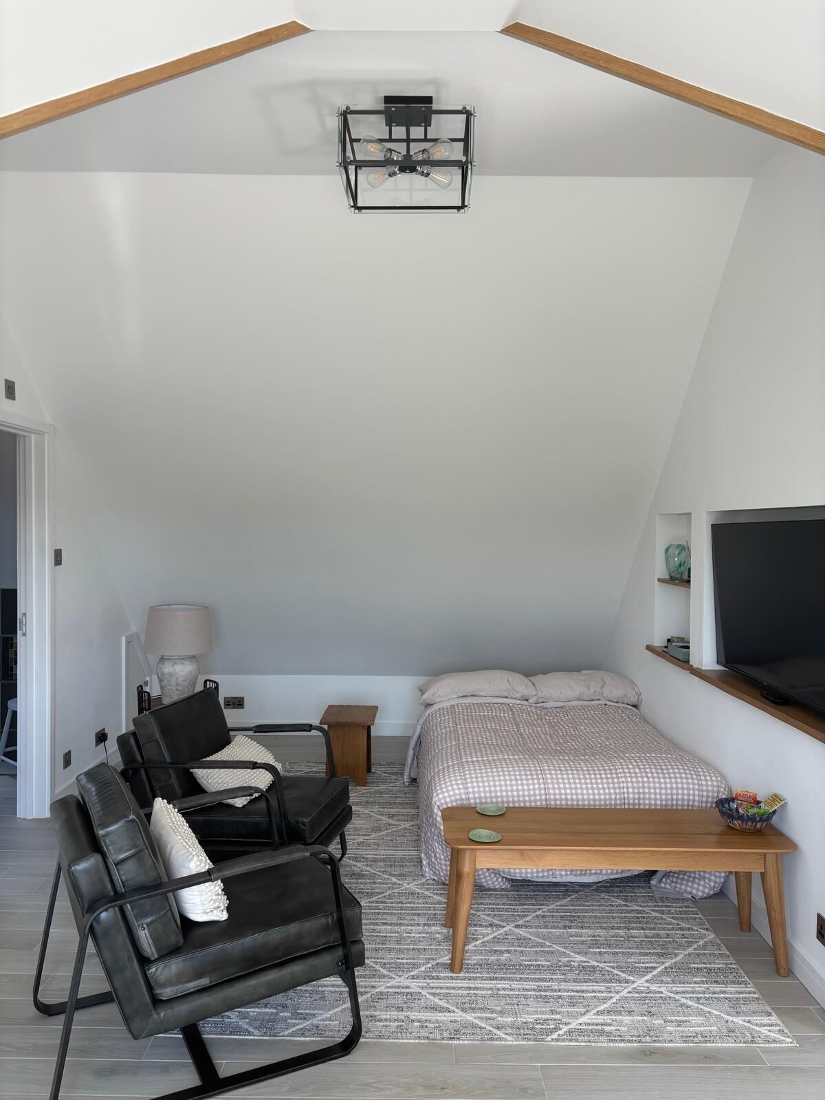A loft bedroom with a sloped ceiling, a bed with a checkered bedspread, a black leather chair, a small wooden table, a floor lamp, and a mounted TV.