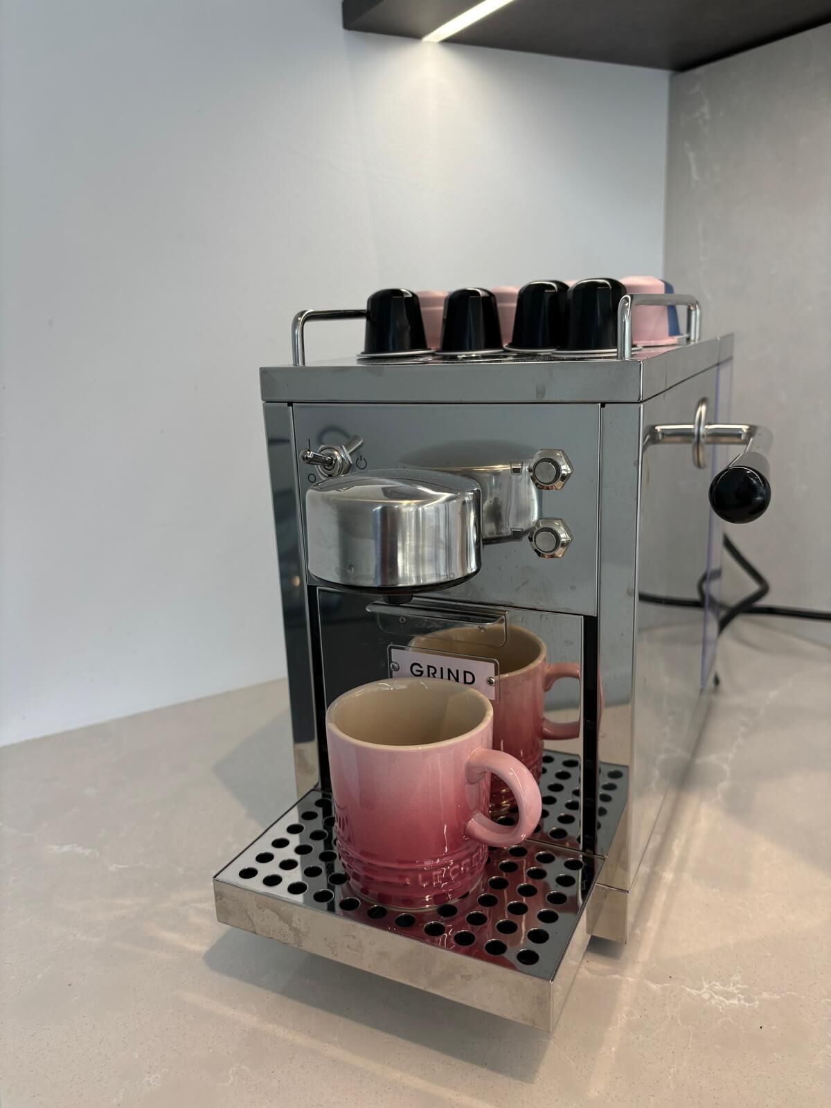 A coffee machine with a single pink mug labeled "GRIND" on a metal tray.