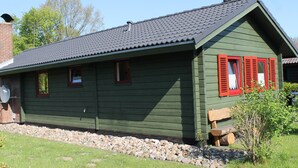 A green wooden building with red shutters, a tiled roof, and a small garden area with a wooden bench and a log. - Cozy log cabin for up to 4 people, free WLAN, fenced property (Damp)