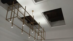 A room with a geometric wooden ceiling structure, recessed lighting, a large window with white curtains, and a door with a light switch. - Dimora Neapolis (Naples)