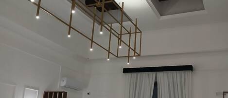 Ein Zimmer mit einer geometrischen Holzdeckenkonstruktion, eingelassener Beleuchtung, einem großen Fenster mit weißen Vorhängen und einer Tür mit Lichtschalter.