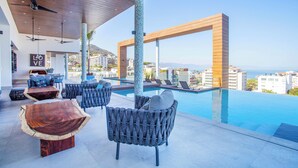A modern outdoor lounge area with a pool, wooden furniture, and a view of the city. - Rooftop Pool + Gym | Romantic Zone Gem (Puerto Vallarta)