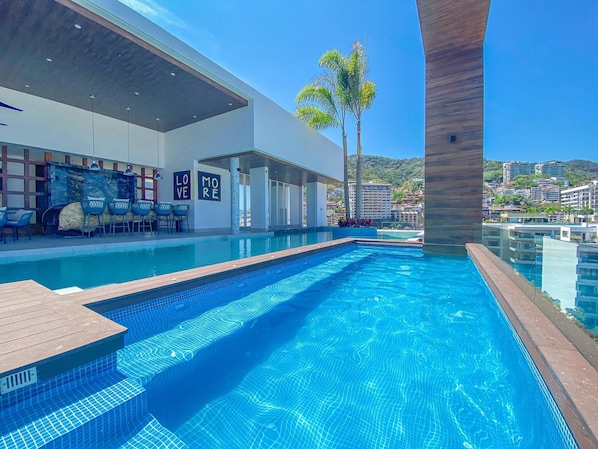 A swimming pool with a wooden deck, a covered seating area, and a view of a cityscape.