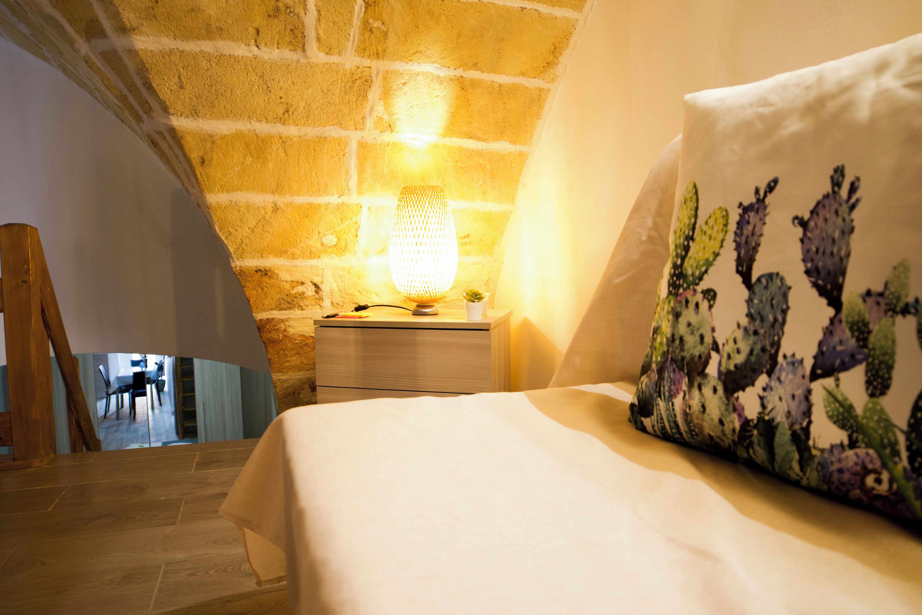 Ein Schlafzimmer mit Backsteinwand, Nachttisch mit Lampe und Bett mit Kaktus-Muster Kissen.
