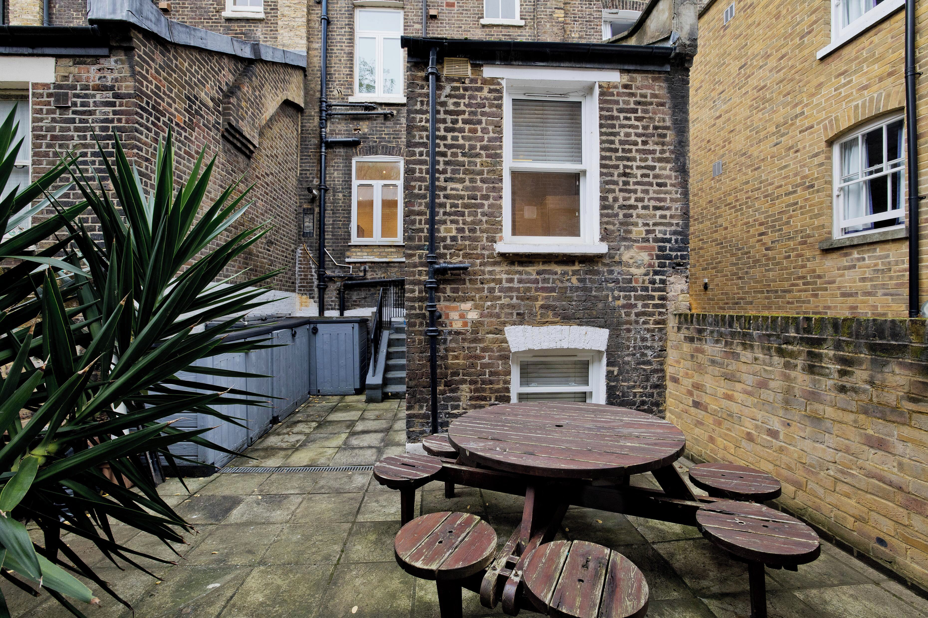 A courtyard with a round wooden table and benches, surrounded by brick walls and a large plant.
