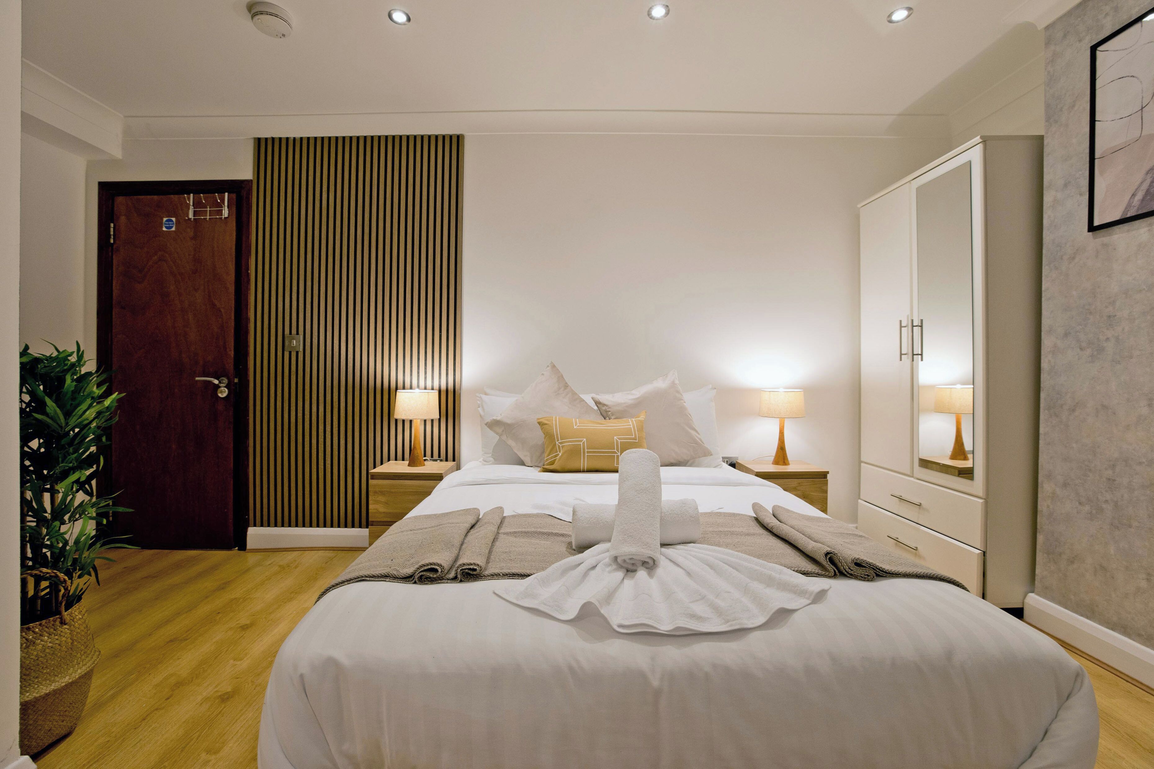 A hotel room with a neatly made bed, a large mirror, a wardrobe, and a bedside table with lamps.