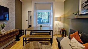 A modern living room with a wooden dining table, a sofa, a TV mounted on the wall, and a large window providing a view of the outdoors. - Imperial Argyle Square Apartment (London)