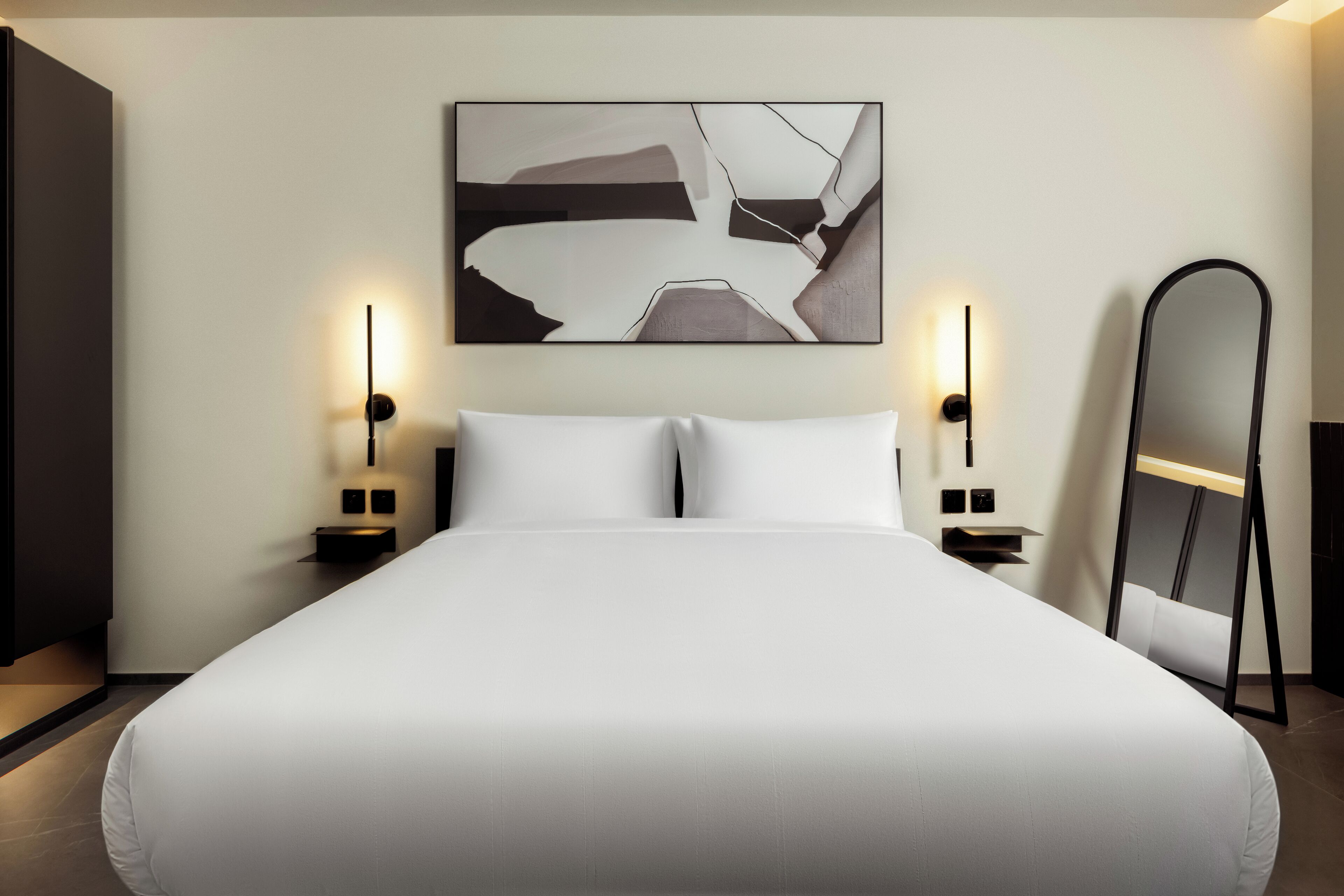 Una habitación de hotel moderna con una cama grande, dos mesitas de noche, un cuadro abstracto colgado en la pared y un espejo de cuerpo entero.