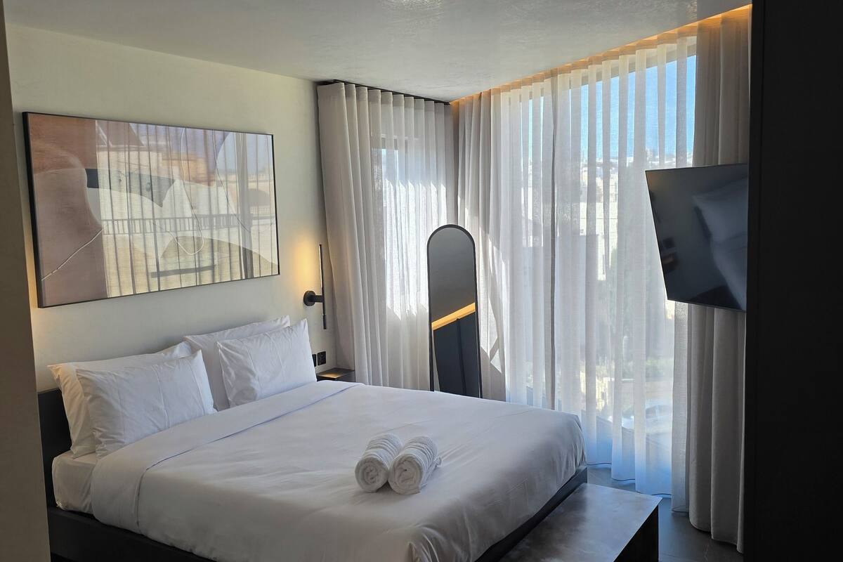 Una habitación de hotel moderna con una cama grande, un televisor de pantalla plana, un escritorio y vistas a la ciudad a través de cortinas translúcidas.