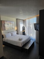 Una habitación de hotel moderna con una cama grande, un televisor de pantalla plana, un escritorio y vistas a la ciudad a través de cortinas translúcidas.