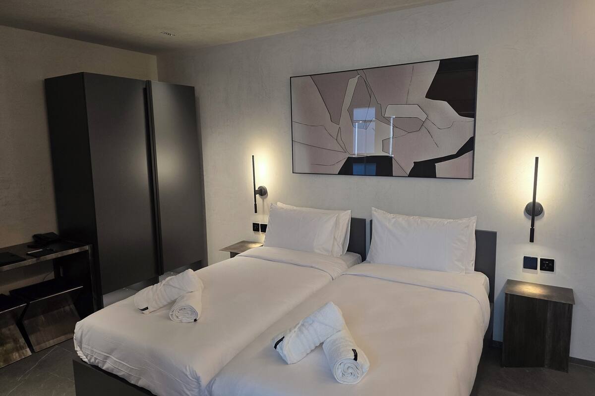 Una habitación de hotel moderna con una cama grande, dos mesitas de noche, un escritorio y una obra de arte abstracta colgada en la pared.