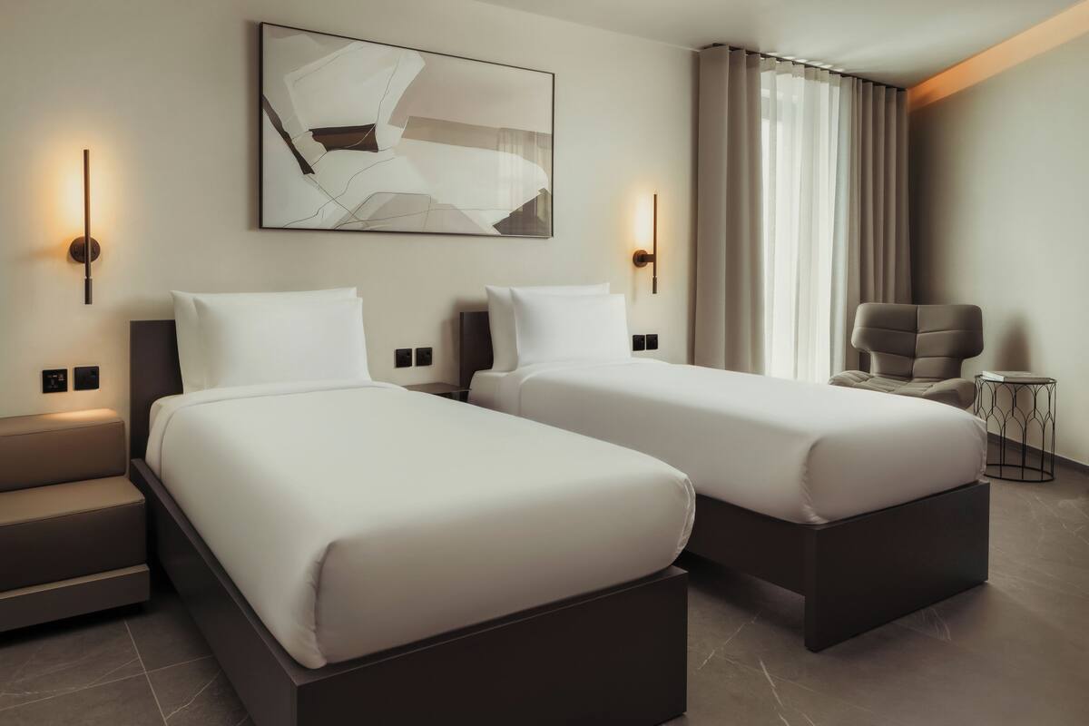 Habitación de hotel con dos camas, una silla, una mesita pequeña y un cuadro abstracto colgado en la pared.
