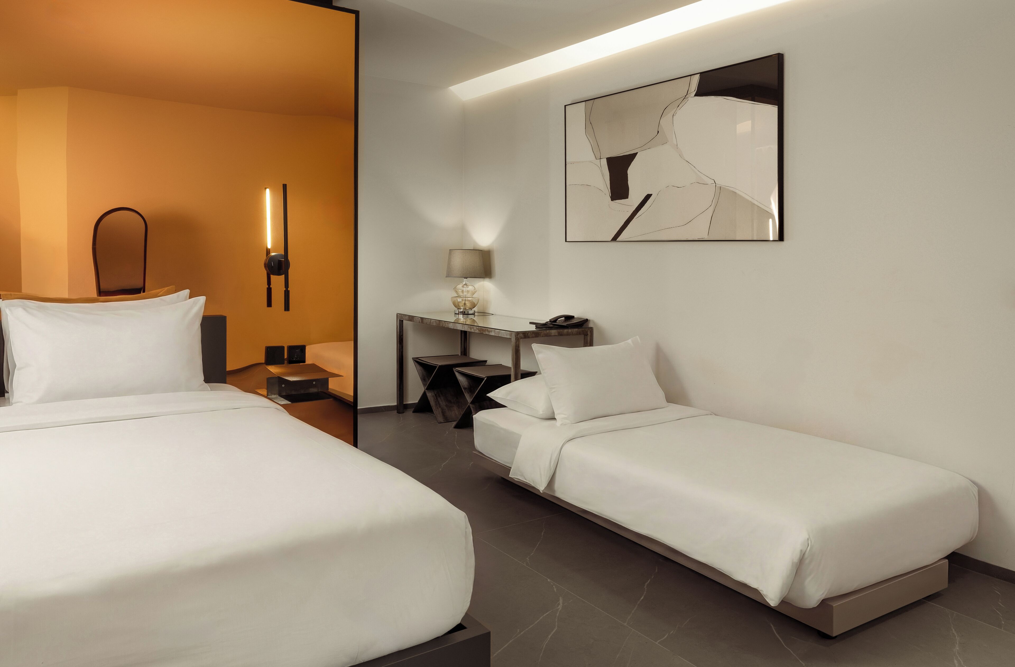 A hotel room with two beds, a desk, a chair, a lamp, and a wall-mounted artwork.