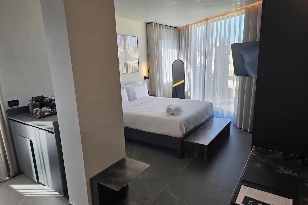 Una habitación de hotel moderna con una cama bien hecha, una mesita de noche pequeña con cafetera y un televisor.