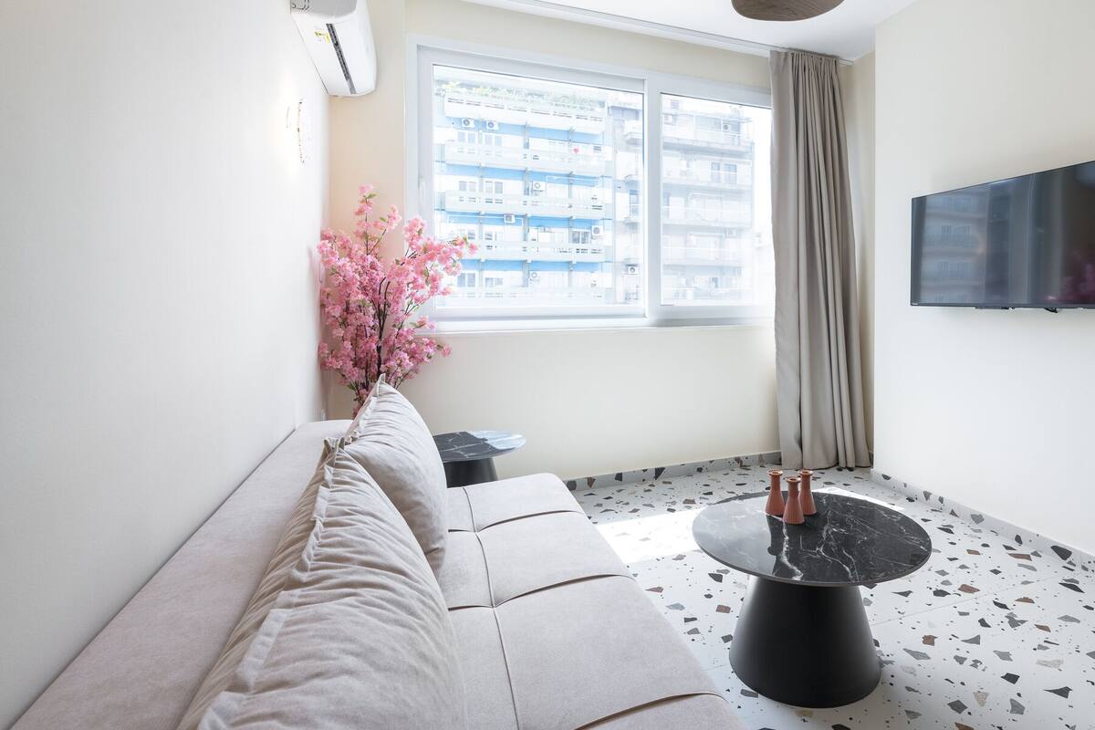 A modern living room with a sofa, a round coffee table, a TV mounted on the wall, and a window with a view of buildings.