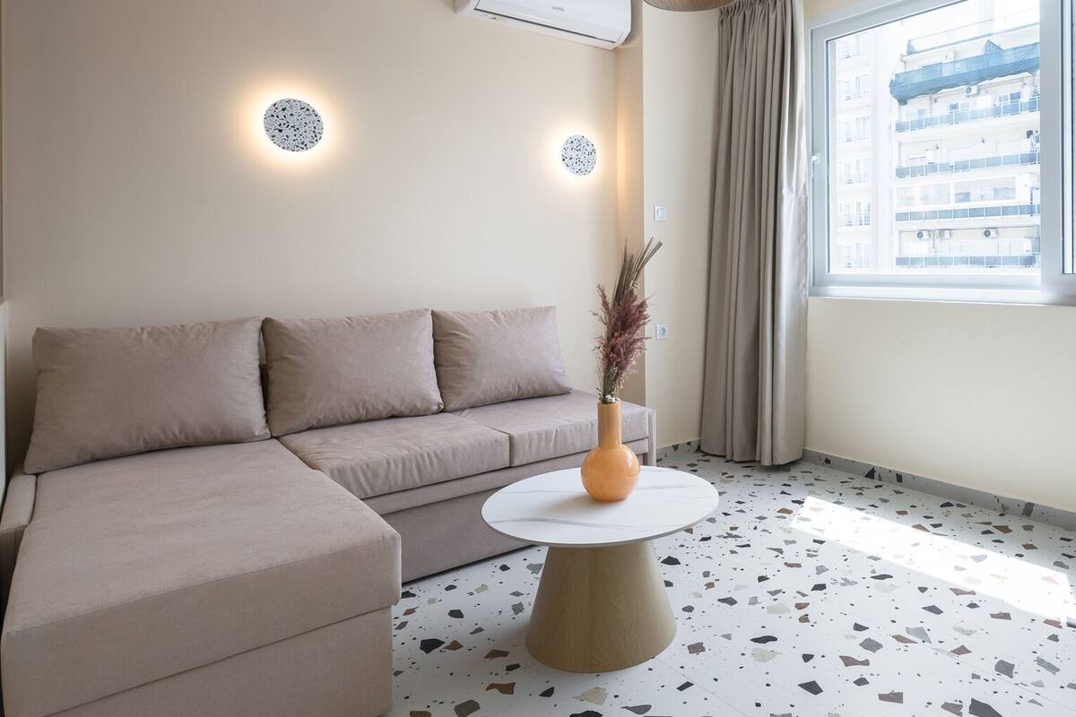 A modern living room with a beige sofa, a round coffee table, and a large window with a city view.