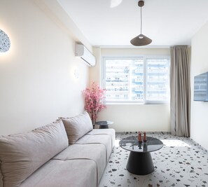 A modern living room with a sofa, a round coffee table, a potted plant, and a large window with a view of buildings.
