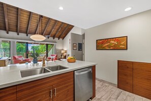 Cucina moderna con soffitto in legno, un'isola grande con lavello e lavastoviglie. C'è un'area pranzo con una sedia rossa e un tavolo, e una finestra che si affaccia sul verde.