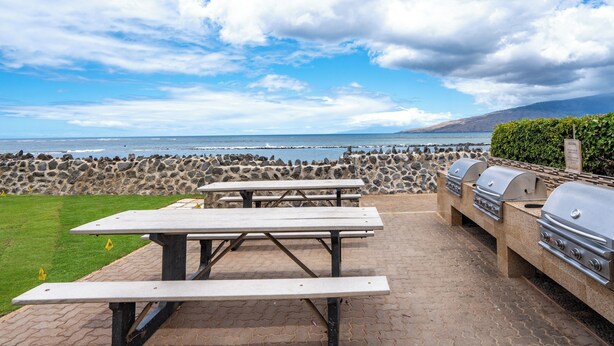 Un patio al aire libre con mesas de picnic y una parrilla, con vista a una costa rocosa y al océano.