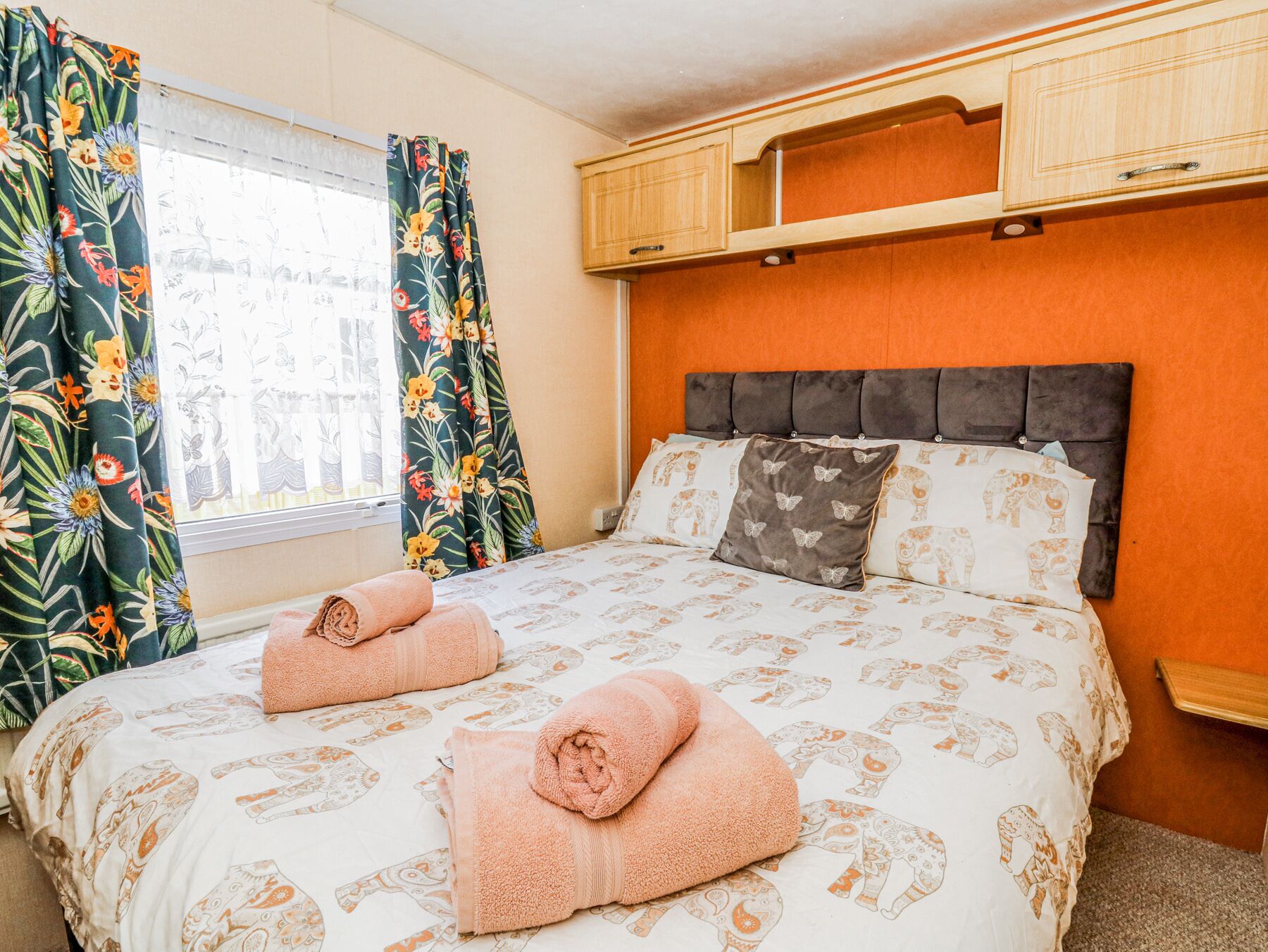 Une chambre d'hôtel avec un lit, deux serviettes enroulées, une fenêtre avec des rideaux à motifs floraux et une armoire en bois au-dessus du lit.