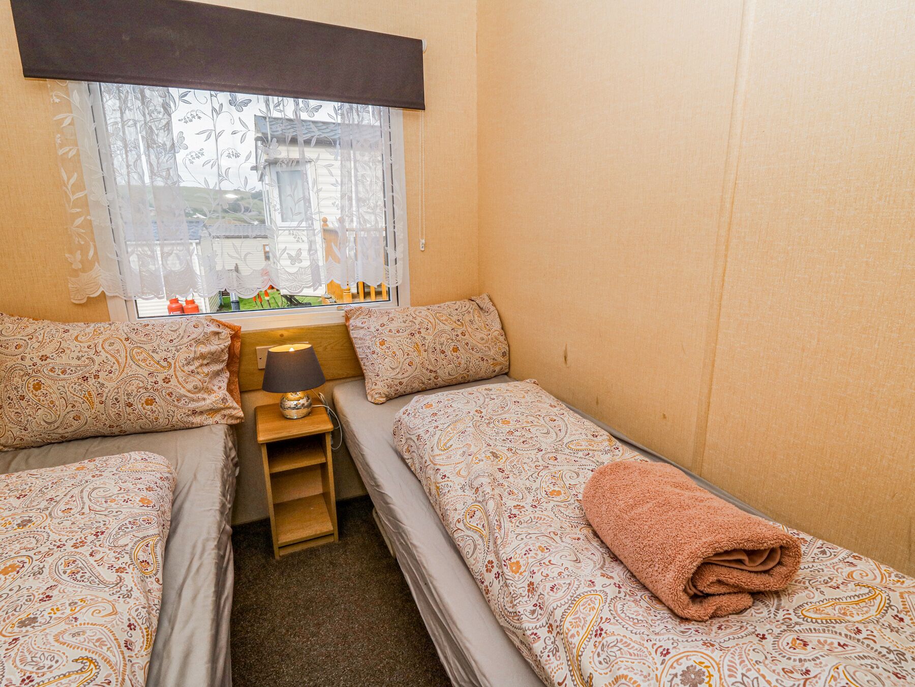 A small, cozy room with two beds, a nightstand, a lamp, and a window with a view of the outside.