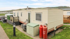 Una casa móvil con una pequeña terraza, un cobertizo verde y dos tanques de propano rojos.