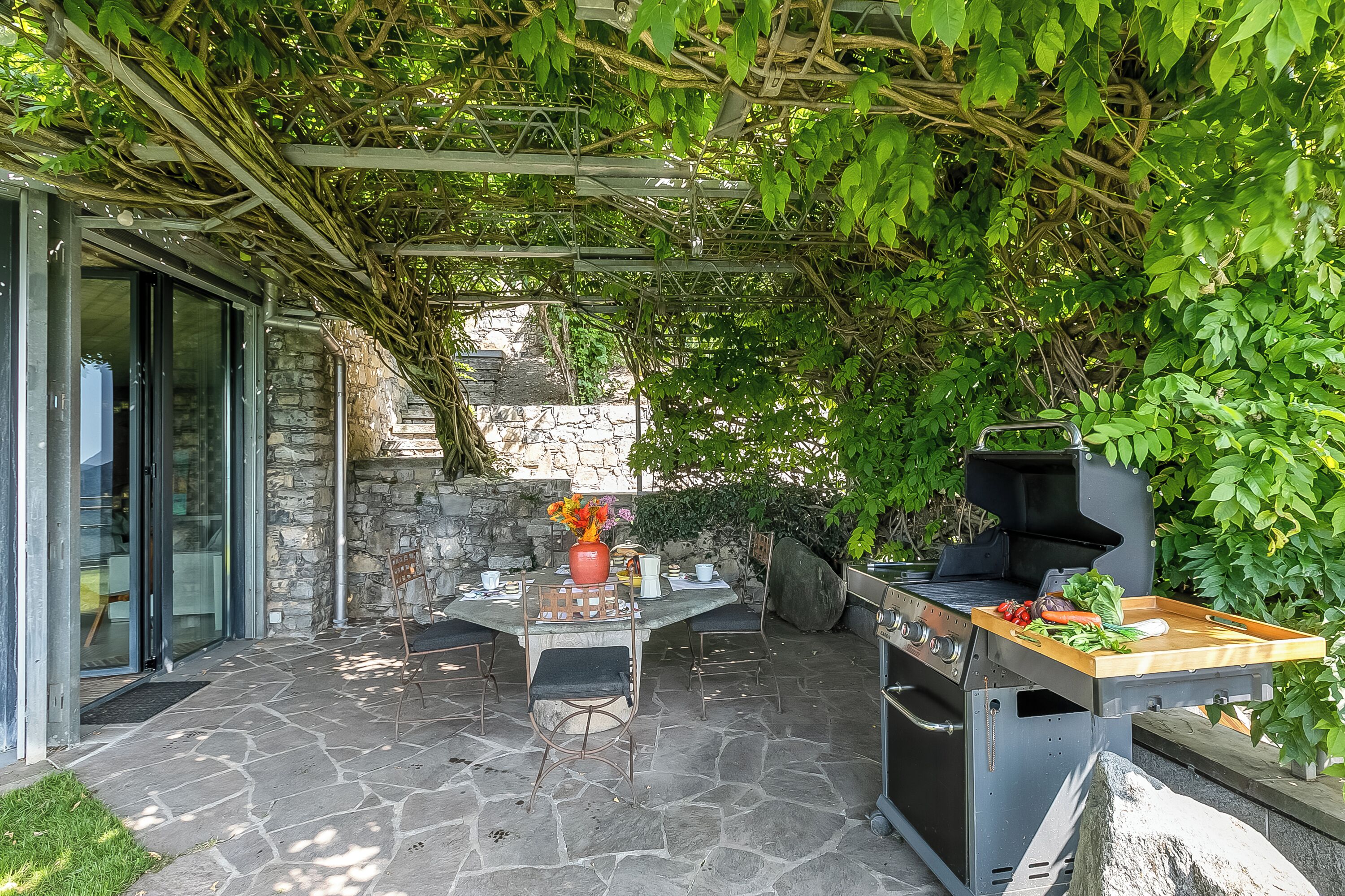 An outdoor patio with stone flooring, a table with chairs, a grill, and a pergola covered with climbing plants.