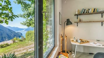 A room with a large window overlooking a mountainous landscape. There is a desk with a lamp, bookshelves, and a tray with books and a cup.