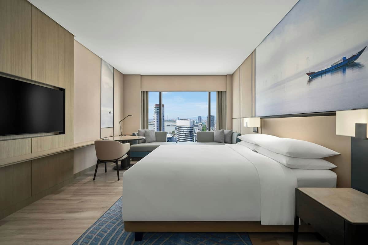 Een moderne hotelkamer met een groot bed, een flatscreen televisie, een zithoek met een stoel en tafel, en een stadszicht door grote ramen.
