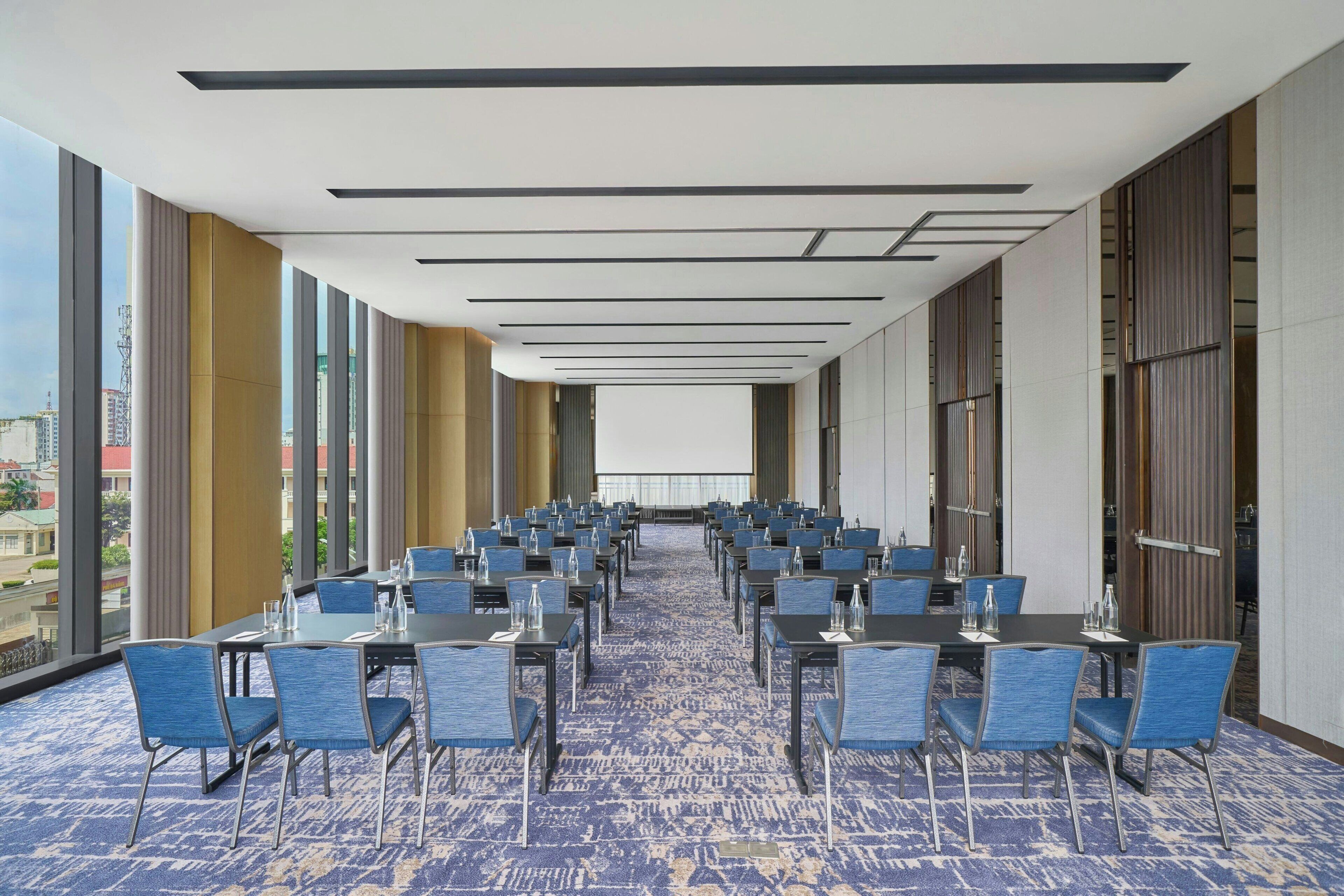 A conference room with rows of tables and chairs, large windows, and a carpeted floor.