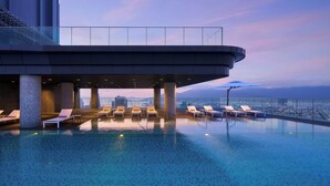 Una piscina infinita en la azotea con sillones de descanso y barandilla de vidrio con vistas a un paisaje urbano.
