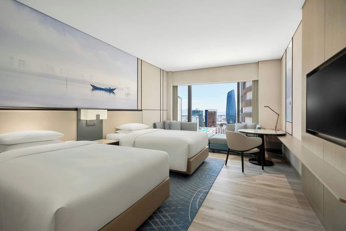 Een hotelkamer met twee bedden, een bureau, een stoel, een televisie en een groot raam met uitzicht op de stad.