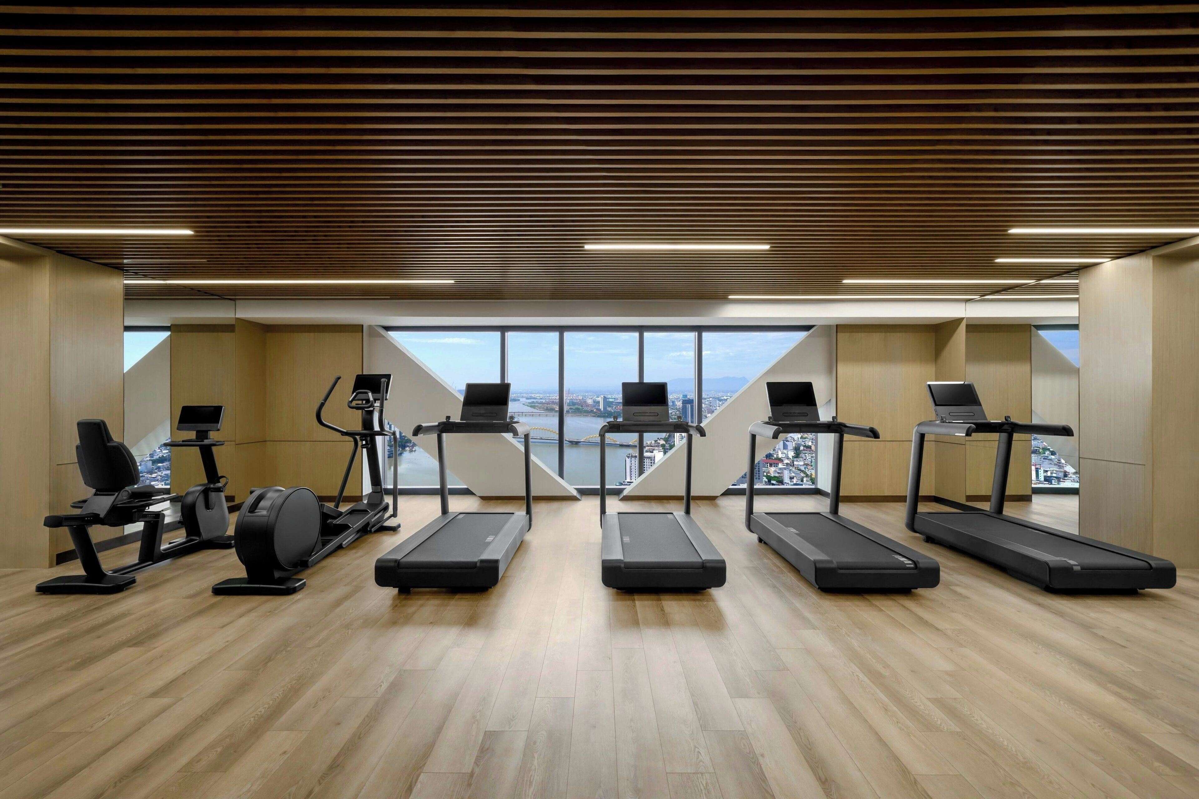 Een fitnessruimte met loopbanden, elliptische trainers en een roeimachine. Er ligt een houten vloer en er is uitzicht op een stadsgezicht door grote ramen.